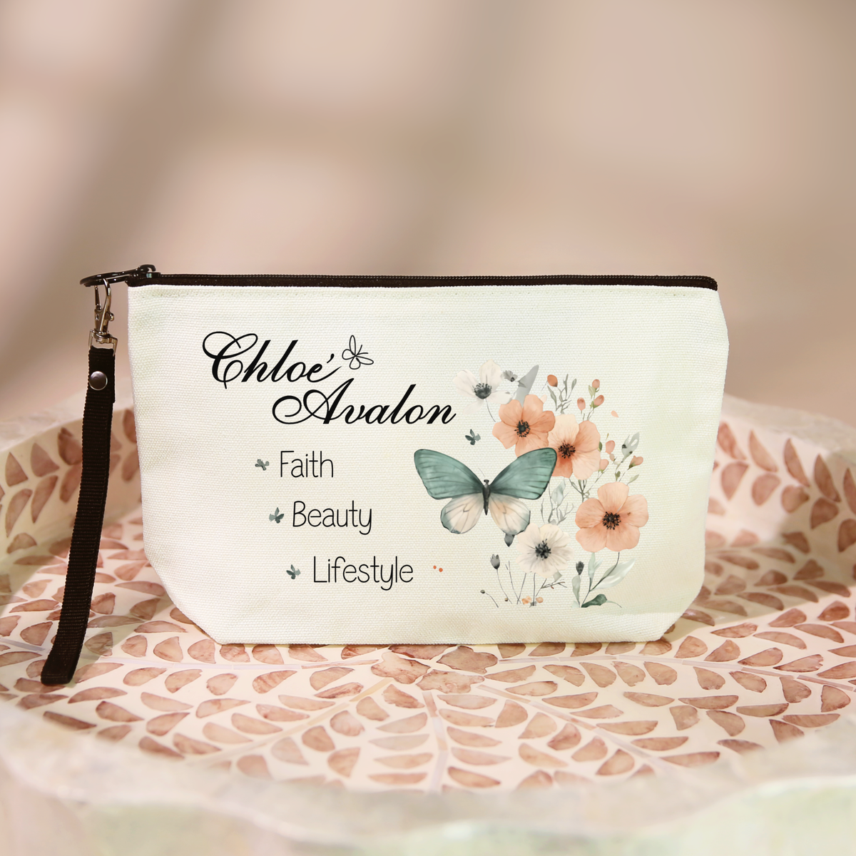 Chloé Avalon cosmetic bag in cream with a black zipper and black wristlet lanyard, printed with the Chloé Avalon script logo, Faith · Beauty · Lifestyle, and a watercolor butterfly with peach florals
